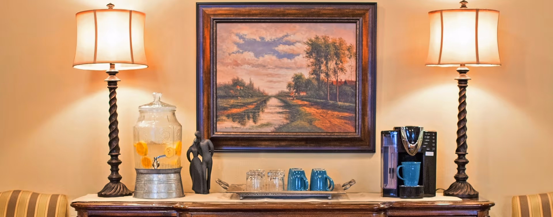 A wooden sideboard with two lit table lamps on either end, a glass beverage dispenser with lemon slices, a small black sculpture of two abstract human figures, a tray with four glasses and two blue mugs, and a coffee maker with a blue mug underneath. Above the sideboard hangs a framed painting of a rural landscape with a dirt path, trees, and a cloudy sky.