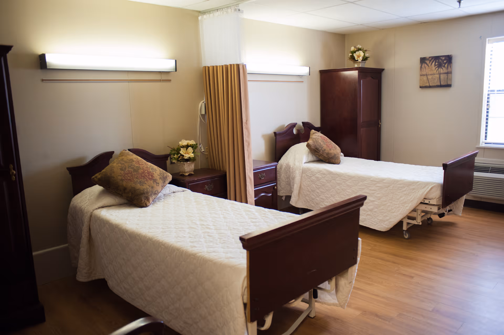 A room with two single beds each covered with white quilted bedspreads and floral pillows. Between the beds is a small wooden nightstand with a flower arrangement. There is a wooden wardrobe and a window with blinds on the right side. The floor is wooden, and the walls are beige. A curtain is partially drawn between the beds.