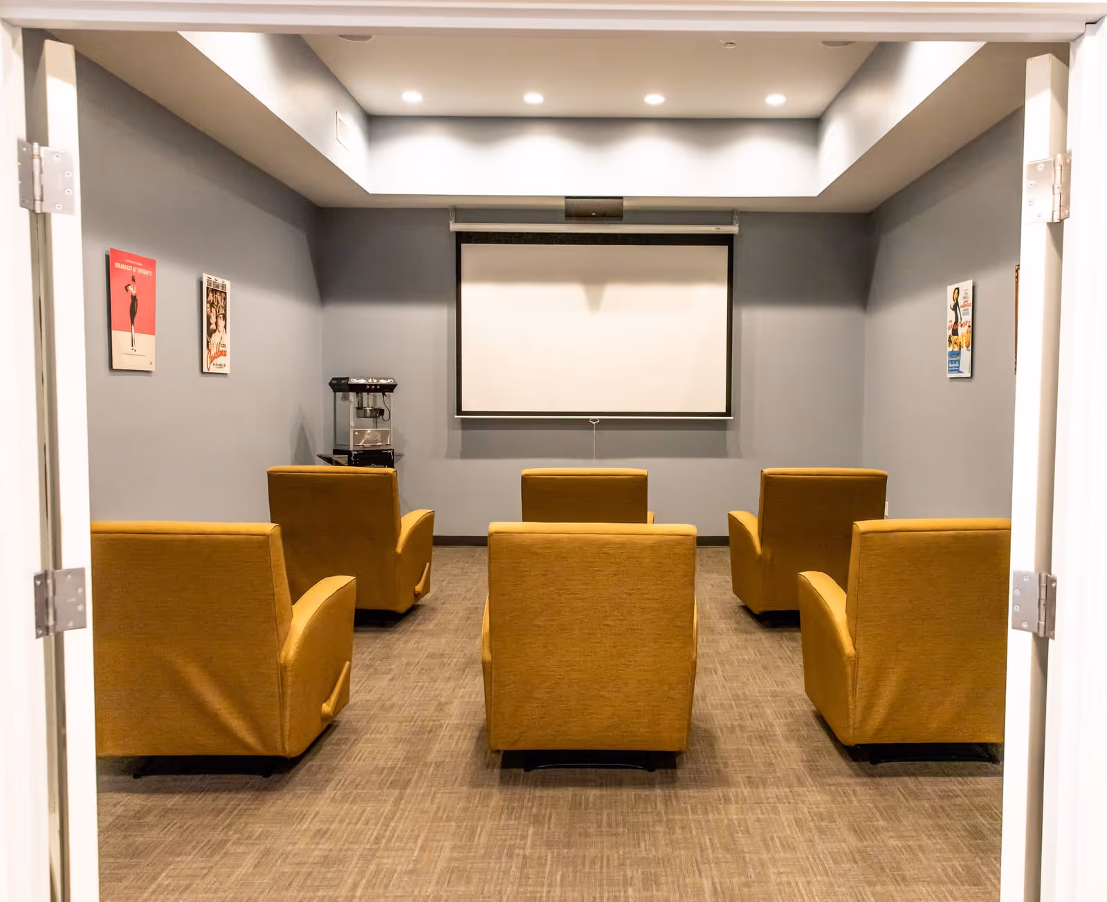 A small theater room with five mustard yellow armchairs arranged in two rows facing a blank projection screen on a gray wall. There is a popcorn machine on a small table to the left side of the screen, and three vintage movie posters are hung on the side walls. The room has a recessed ceiling with soft lighting.