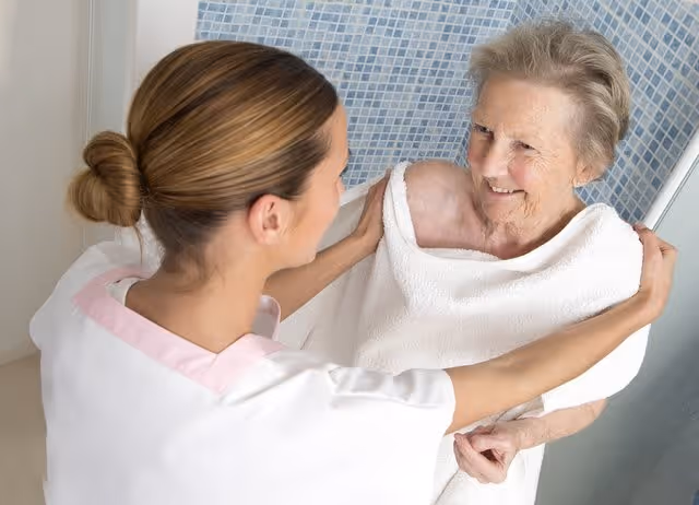 A caregiver with her hair in a bun is wrapping a white towel around an elderly woman who is smiling, standing in front of a tiled shower area.