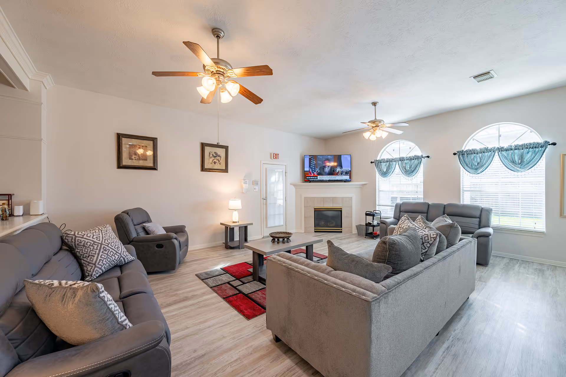 A bright and spacious living room in Serenity Pointe Assisted Living featuring multiple gray sofas and armchairs with patterned cushions, a coffee table on a red and gray area rug, two ceiling fans with lights, a fireplace with a mounted flat-screen TV above it, and large arched windows with blue curtains allowing natural light to fill the room.