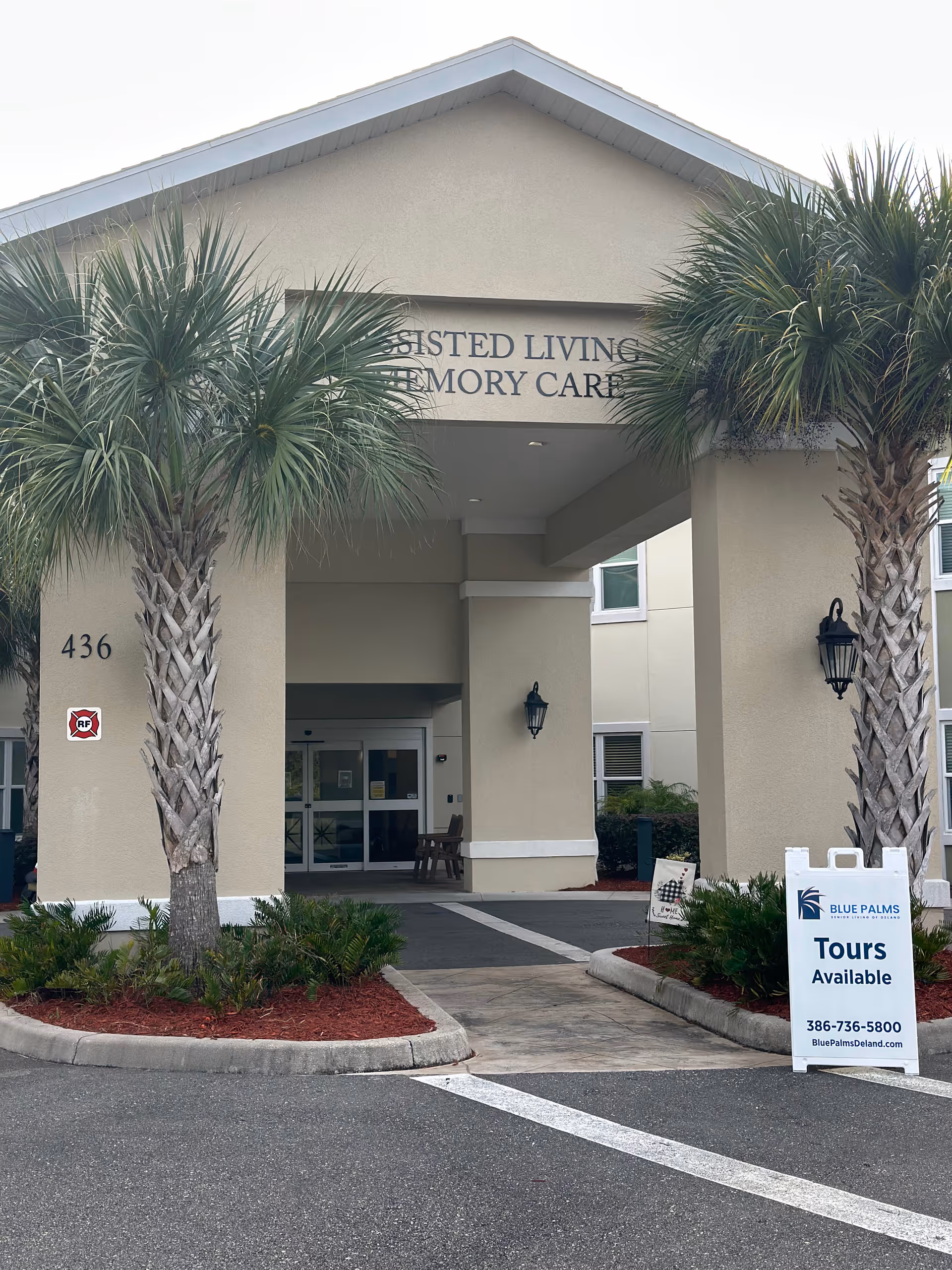 Entrance of a senior living facility with a covered driveway flanked by two palm trees. The building has beige walls and a sign above the entrance partially visible reading 'Assisted Living Memory Care'. There is a white sandwich board sign on the right side advertising tours available with contact information for Blue Palms Senior Living of Deland.