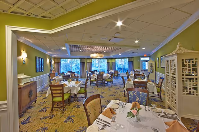 A spacious dining room with multiple tables set for a meal, featuring white tablecloths, folded napkins, glassware, and floral centerpieces. The room has green walls, large windows with curtains, decorative lighting fixtures, and a patterned carpet. Wooden chairs with cushioned seats surround the tables.