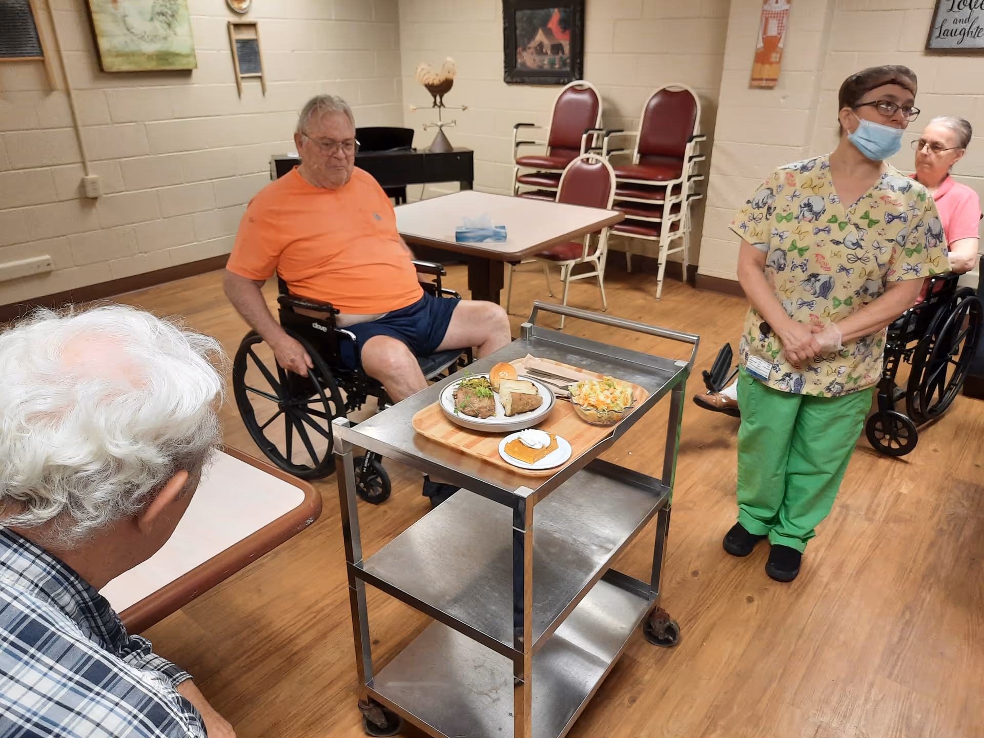 Elderly residents and a masked staff member standing around a meal cart in a communal dining/activity room.