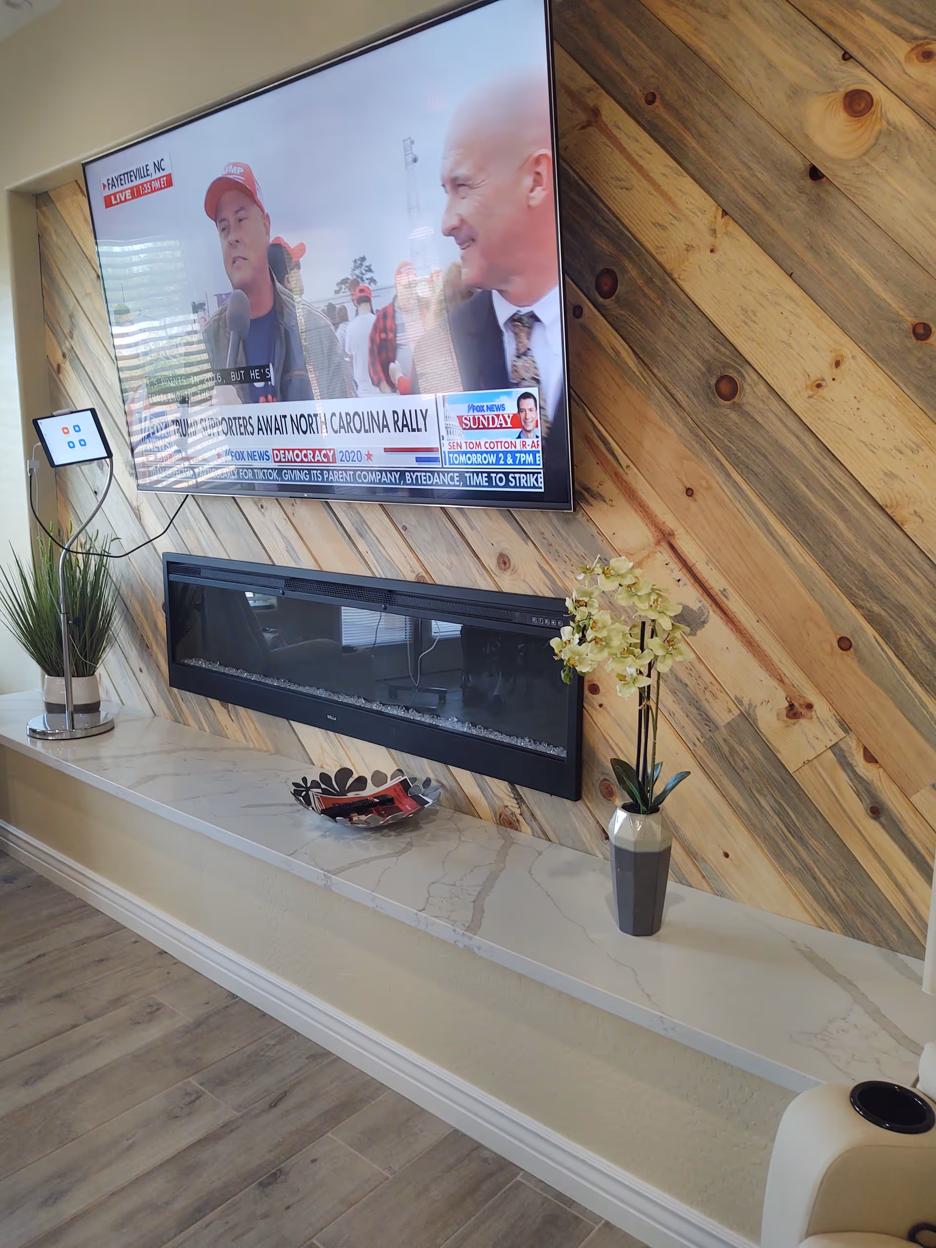 A modern living room area featuring a wall-mounted flat screen TV displaying a news broadcast, a sleek black electric fireplace below it, and a long marble shelf beneath the fireplace. The wall behind the TV and fireplace is decorated with diagonal wooden panels. On the shelf, there is a decorative bowl and a vase with white flowers. A potted plant is placed on the left side of the shelf.