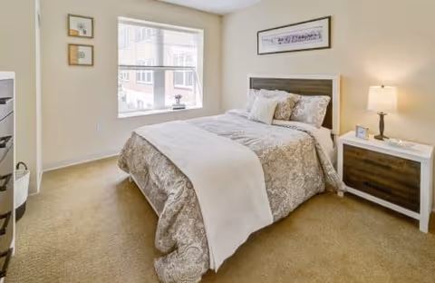 Well-lit bedroom with a neatly made double bed, nightstand with lamp, window and framed wall art.