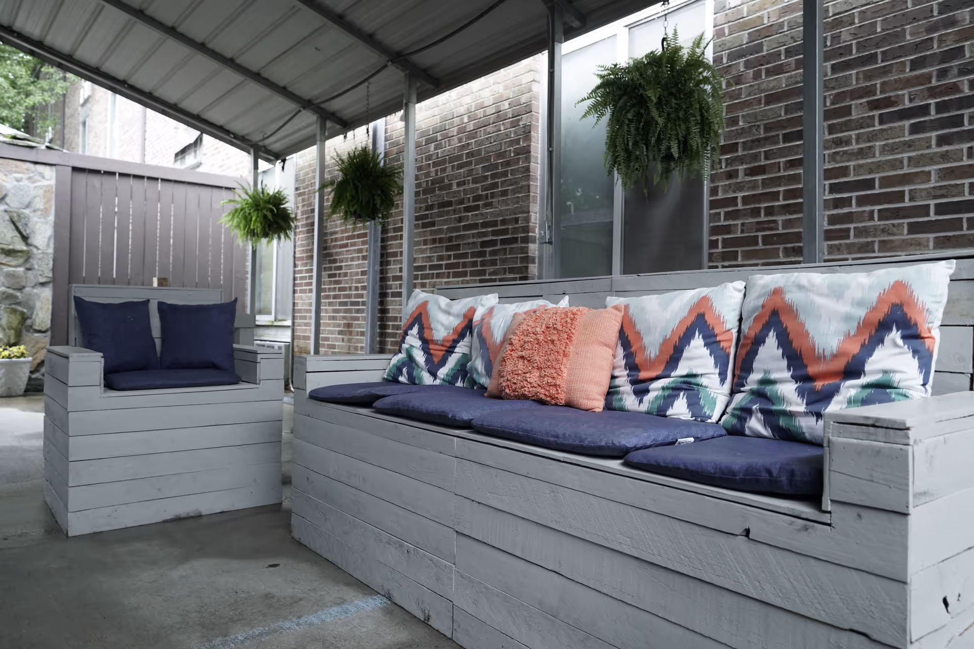 Covered outdoor seating area with a wooden bench and chair painted in light gray, adorned with colorful cushions including zigzag patterned pillows and solid color pillows. Hanging green plants are suspended from the ceiling, and a brick wall with windows is visible in the background.
