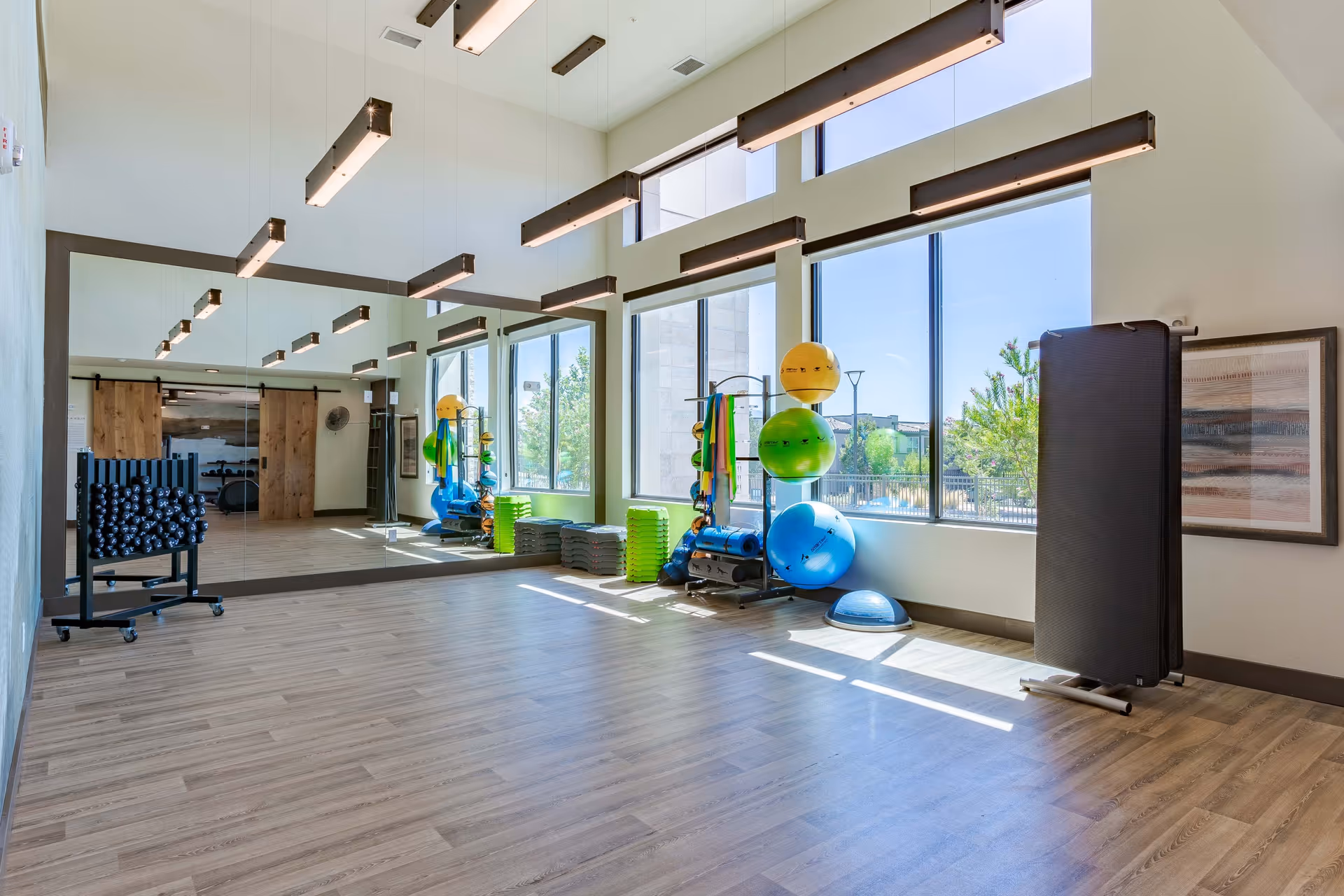 Bright exercise room with large windows letting in natural light, wooden floor, wall-length mirror, and various fitness equipment including exercise balls, step platforms, and weights.