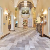Spacious and well-lit interior hallway with tiled floor, arched doorways, and warm beige walls. There is a wooden reception desk on the right side, decorative wall sconces, and a round table with a floral arrangement in the center. A balcony with railing is visible above the far end of the hallway.