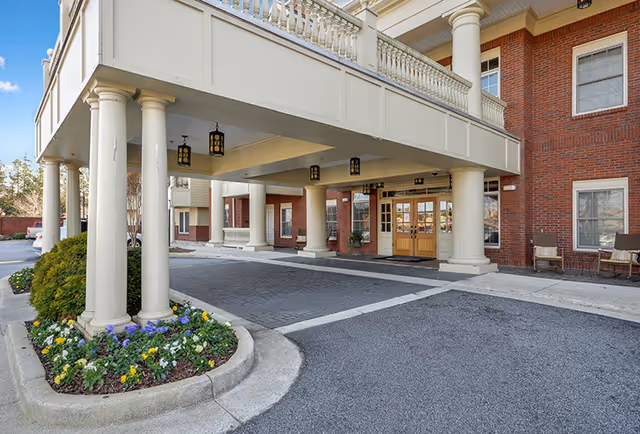 Entrance area of a senior living facility with a covered driveway supported by large white columns. The building has red brick walls and large wooden double doors. There are flower beds with colorful flowers and some outdoor chairs near the entrance.
