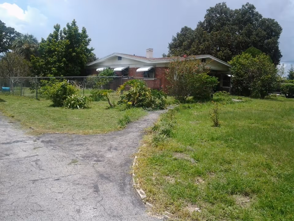 A single-story brick house with white awnings over the windows, surrounded by a grassy yard with various bushes and trees. A paved driveway curves in front of the house, and a chain-link fence encloses part of the yard.