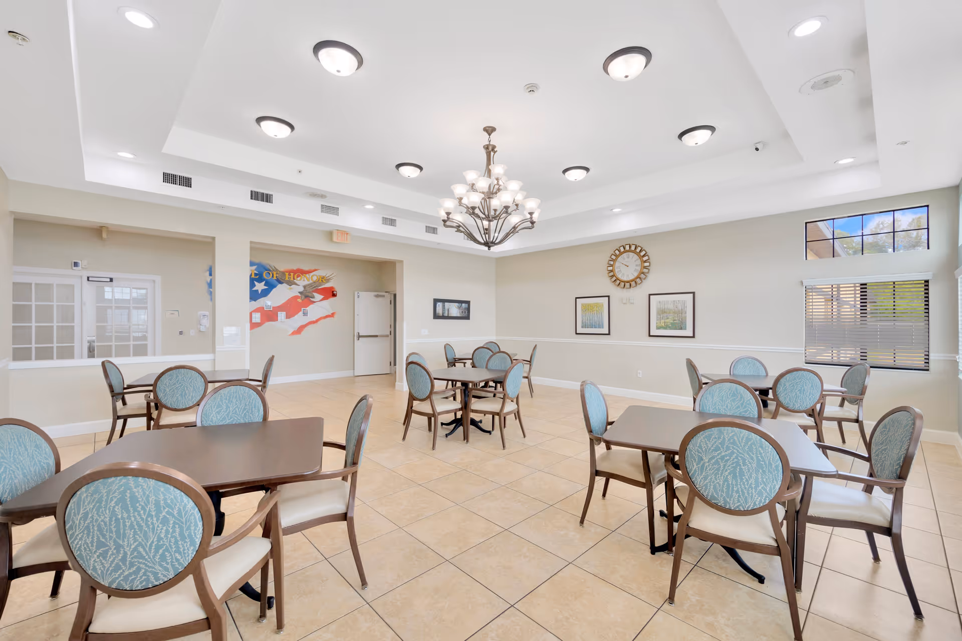 A bright and spacious dining room with multiple square tables and chairs featuring blue patterned upholstery. The room has beige tiled floors, cream-colored walls, and a large chandelier hanging from a recessed ceiling with additional ceiling lights. There are framed pictures and a clock on the wall, and windows with blinds letting in natural light. A mural with an American flag and the words 'Wall of Honor' is visible on one wall.