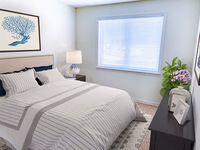 Well-lit bedroom with a striped bed, bedside table and lamp, dresser with decorative items, and a window with closed blinds.