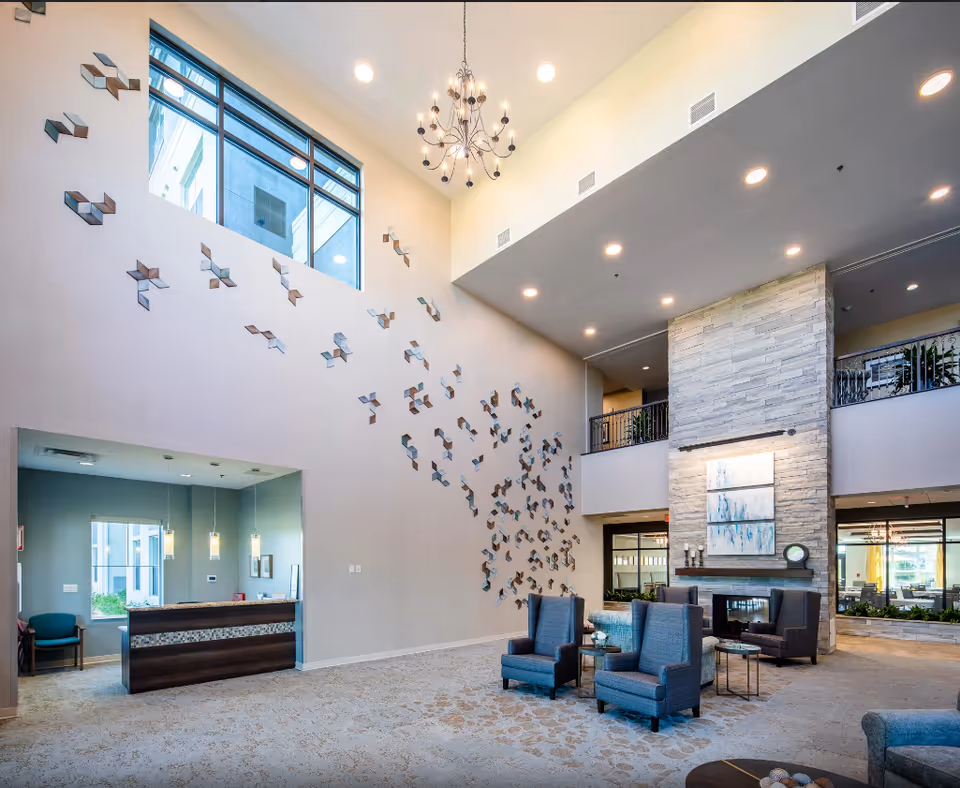 Spacious senior living facility lobby with high ceilings, a modern chandelier, and large windows. The room features a stone fireplace with artwork above it, several blue upholstered armchairs arranged around small tables, and a reception desk area with pendant lighting. Decorative wall art is displayed on the tall wall near the windows.