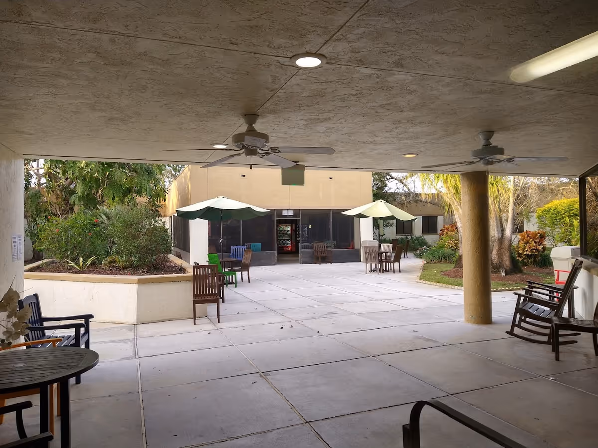 Covered outdoor courtyard with tables, chairs, umbrellas and ceiling fans at a senior living facility.