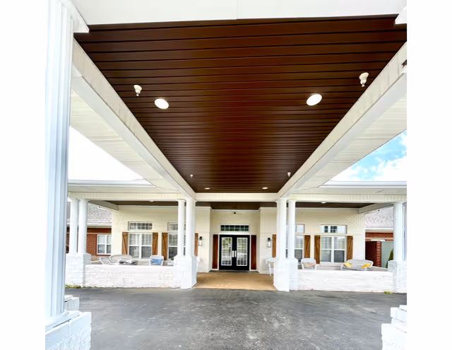 Covered entry/porte-cochere with white columns, seating areas, and double glass doors leading into a senior living building.