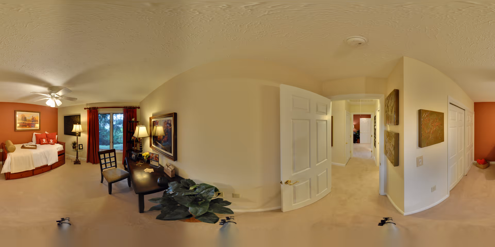 A spacious bedroom with beige walls and carpet, featuring a bed with red and white bedding, a ceiling fan, a window with red curtains overlooking greenery, a desk with a chair and lamp, framed artwork on the walls, and a hallway leading to other rooms.