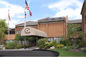 Exterior view of a two-story brick building with a covered entrance canopy displaying a circular logo. There are three flagpoles with flags in front of the building, surrounded by landscaped greenery and a paved driveway.