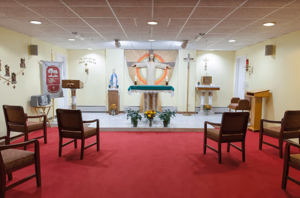 Interior of a small chapel with a red carpeted floor and wooden chairs arranged facing an altar. The altar features a statue of Jesus with outstretched arms, flanked by religious symbols including a crucifix, a statue of the Virgin Mary, candles, and flowers. The walls are light yellow, and there is a banner on the left side with religious imagery and text that reads 'St. Charles Pray For Us'.