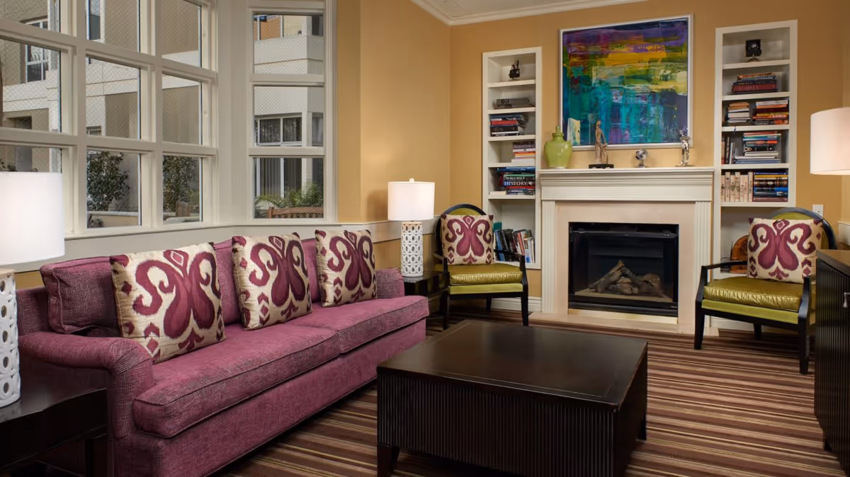 A cozy living room with a purple sofa adorned with patterned cushions, two green armchairs with matching cushions, a dark wooden coffee table, a fireplace with a colorful abstract painting above it, built-in white bookshelves filled with books and decorative items, and two white table lamps on either side of the sofa. Large windows let in natural light and show an exterior view of a building.