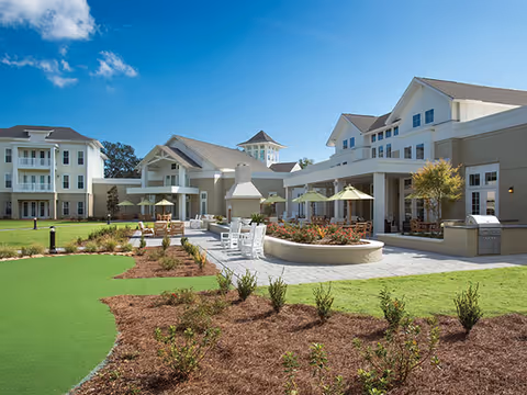Outdoor patio area at South Bay At Mount Pleasant featuring seating with umbrellas, a fireplace, landscaped garden beds, and multi-story residential buildings under a blue sky with scattered clouds.