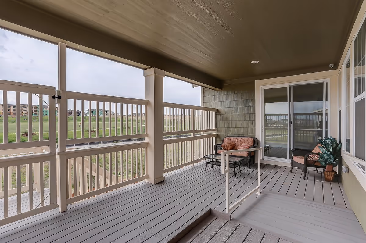 Covered outdoor balcony with gray wooden flooring and beige railing, furnished with cushioned wicker chairs and a small glass-top table, overlooking a grassy area and distant buildings under a cloudy sky.