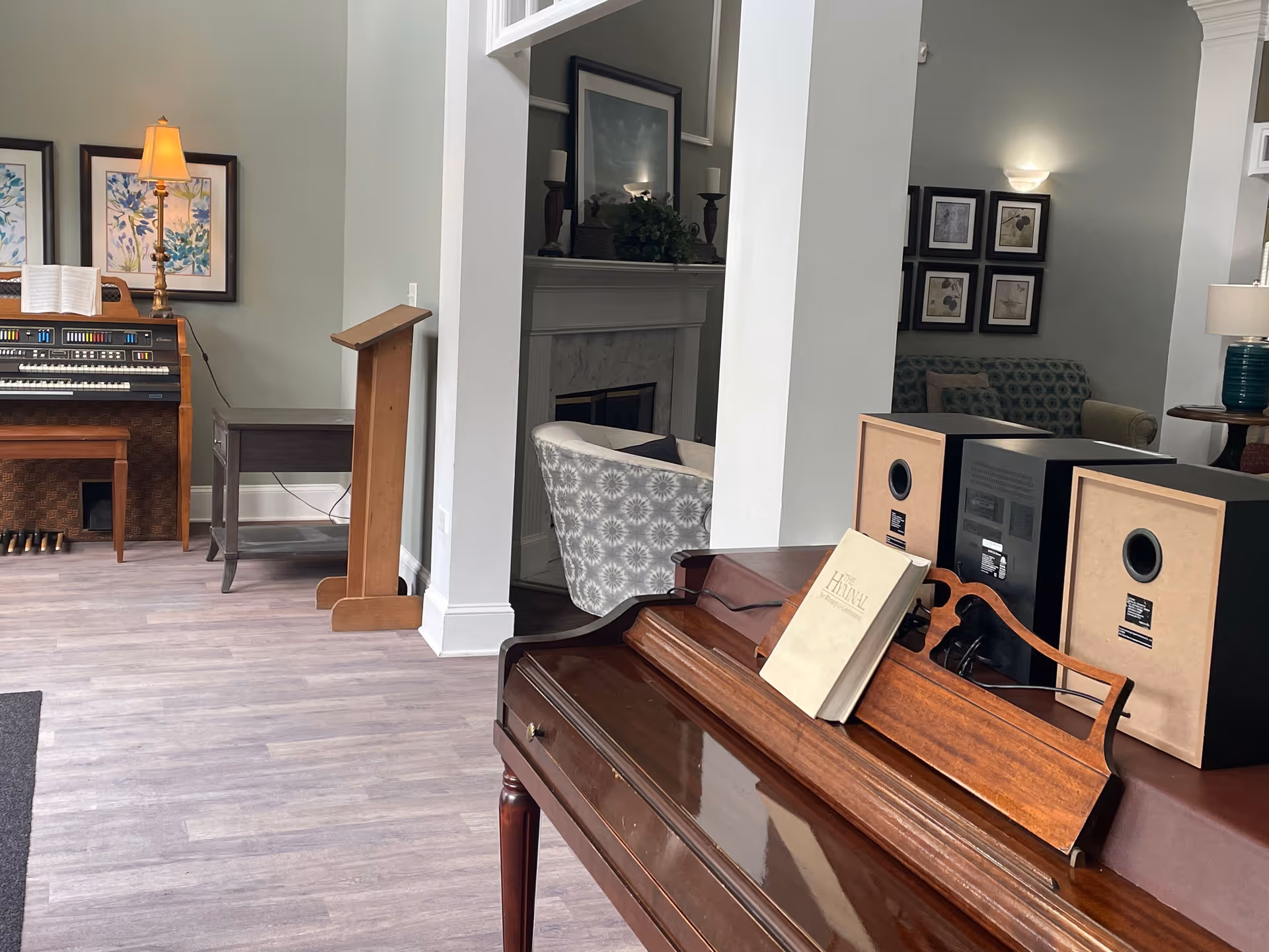 Interior view of a senior living facility common area featuring a wooden piano with a music book titled 'The Hymnal' and two speakers on top. In the background, there is an electric organ with a bench, a wooden music stand, a fireplace with decorative items, and comfortable seating including a patterned armchair and a sofa with framed artwork on the walls.