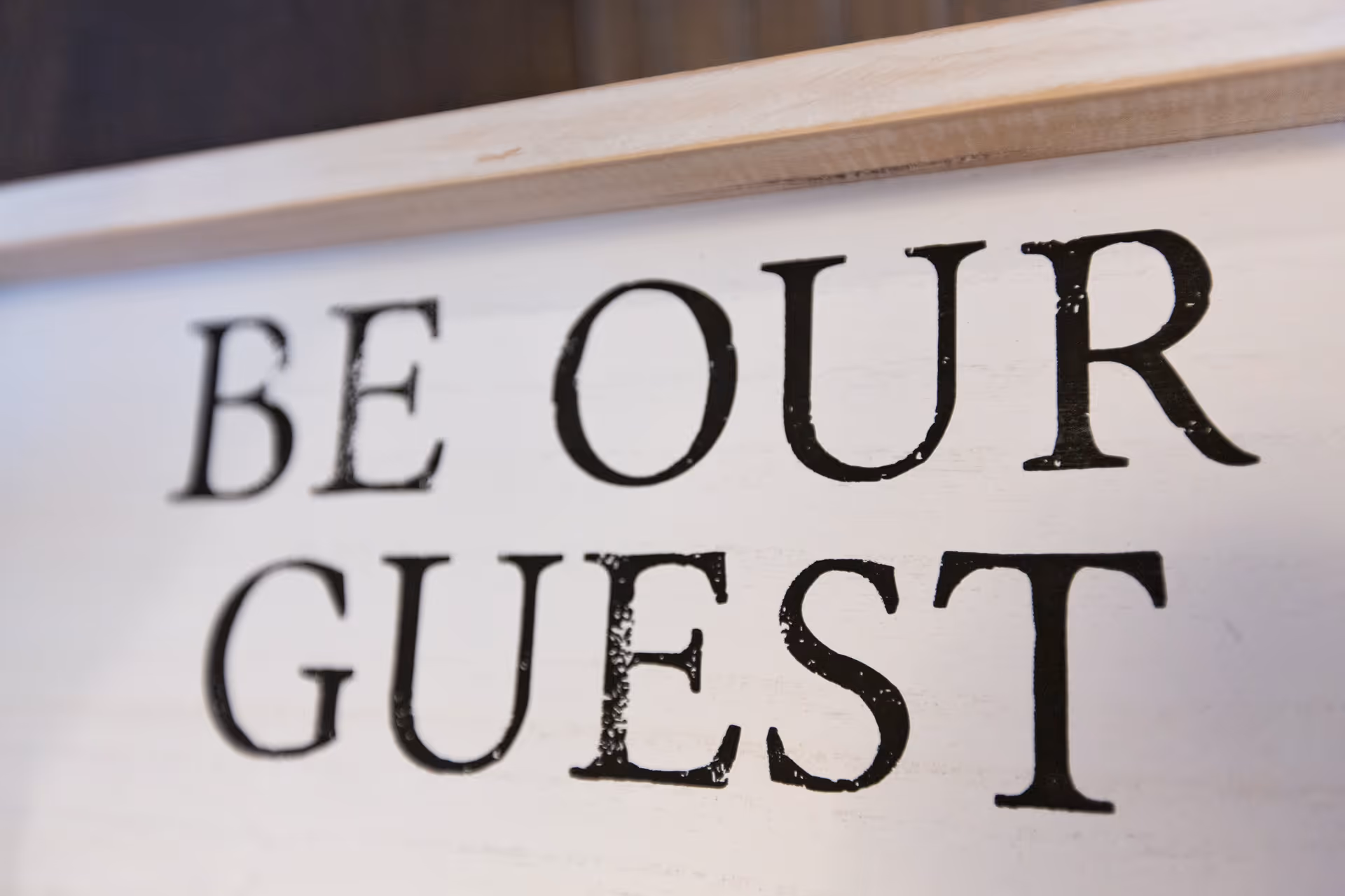 Close-up of a decorative sign with the words 'BE OUR GUEST' written in black letters on a white wooden background.