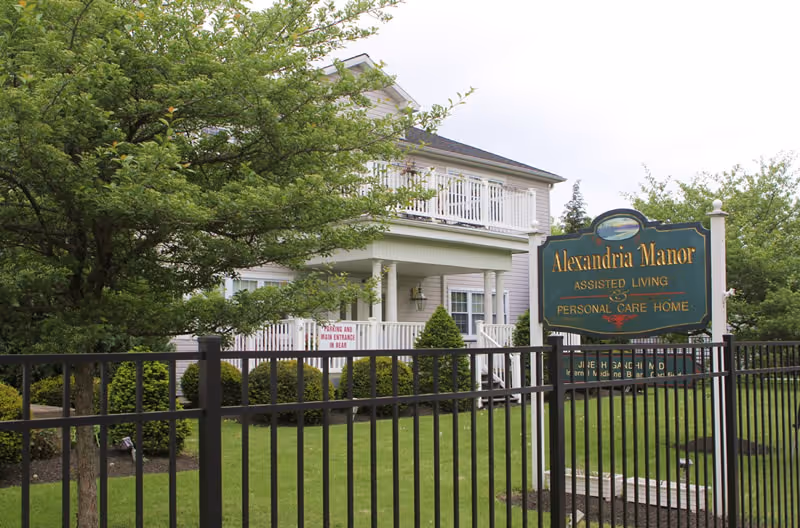 Two-story Alexandria Manor assisted living building with a lawn and entrance sign behind a black metal fence.