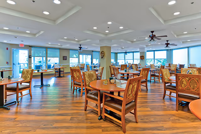 A spacious dining room with multiple round wooden tables and cushioned chairs arranged neatly. The room has large windows with blue blinds, ceiling fans, and recessed lighting. The floor is wooden, and the room is bright with natural light coming through the windows.