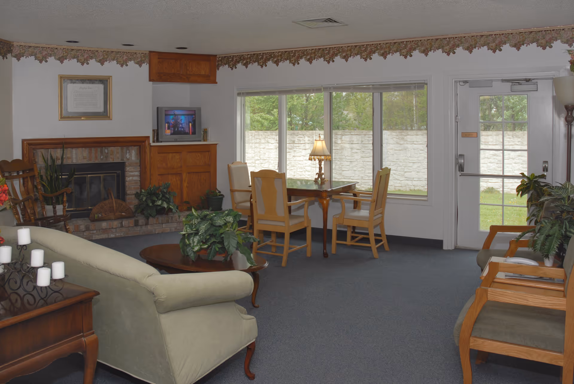 A cozy living room area in a senior living facility with a green upholstered sofa, wooden chairs, a brick fireplace, a small TV on a wooden cabinet, a table with four chairs near large windows, and various plants and lamps providing a warm atmosphere.
