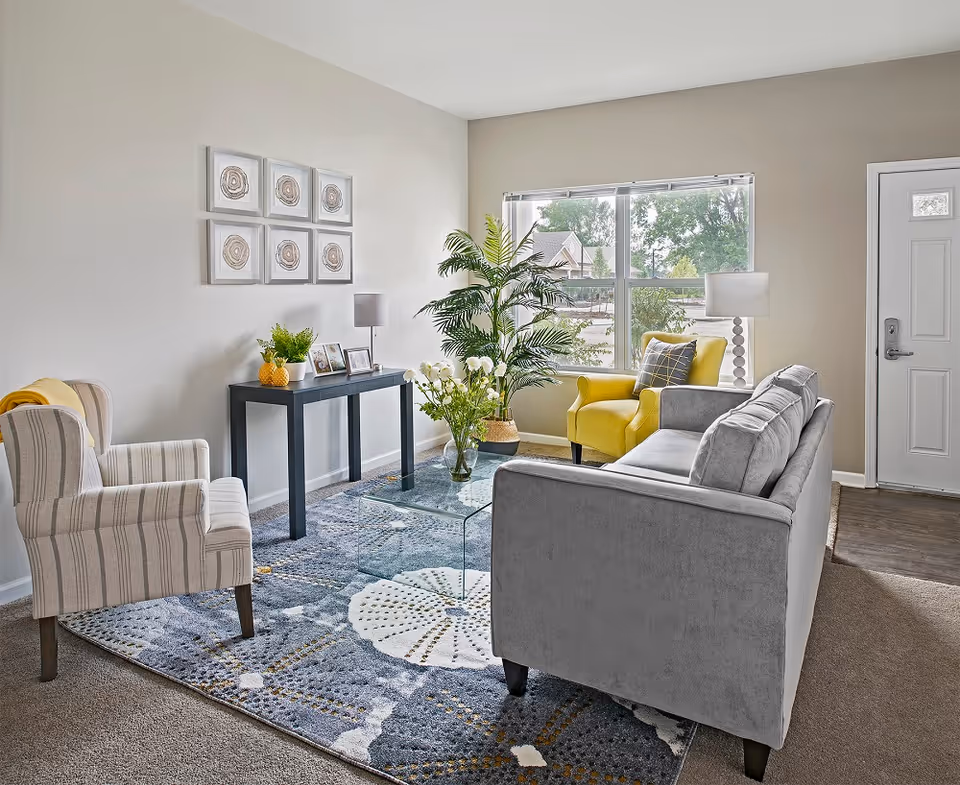 Bright living room with a gray sofa, yellow armchair, striped accent chair, glass coffee table, console table and a large window.