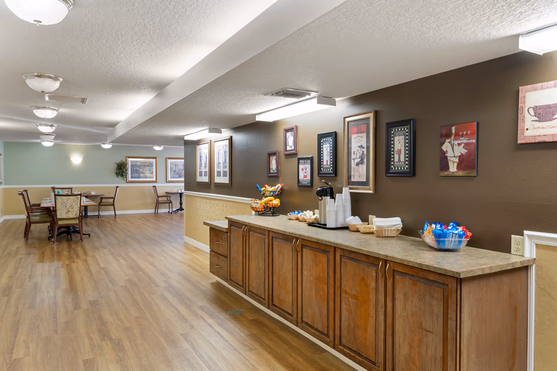 A spacious interior common area with wooden flooring and a long countertop on the right side. The countertop has baskets with snacks, a coffee dispenser with cups, and napkins. The wall above the countertop is decorated with framed pictures. In the background, there are tables and chairs arranged for dining or socializing, with framed artwork on the walls and soft lighting from ceiling fixtures.