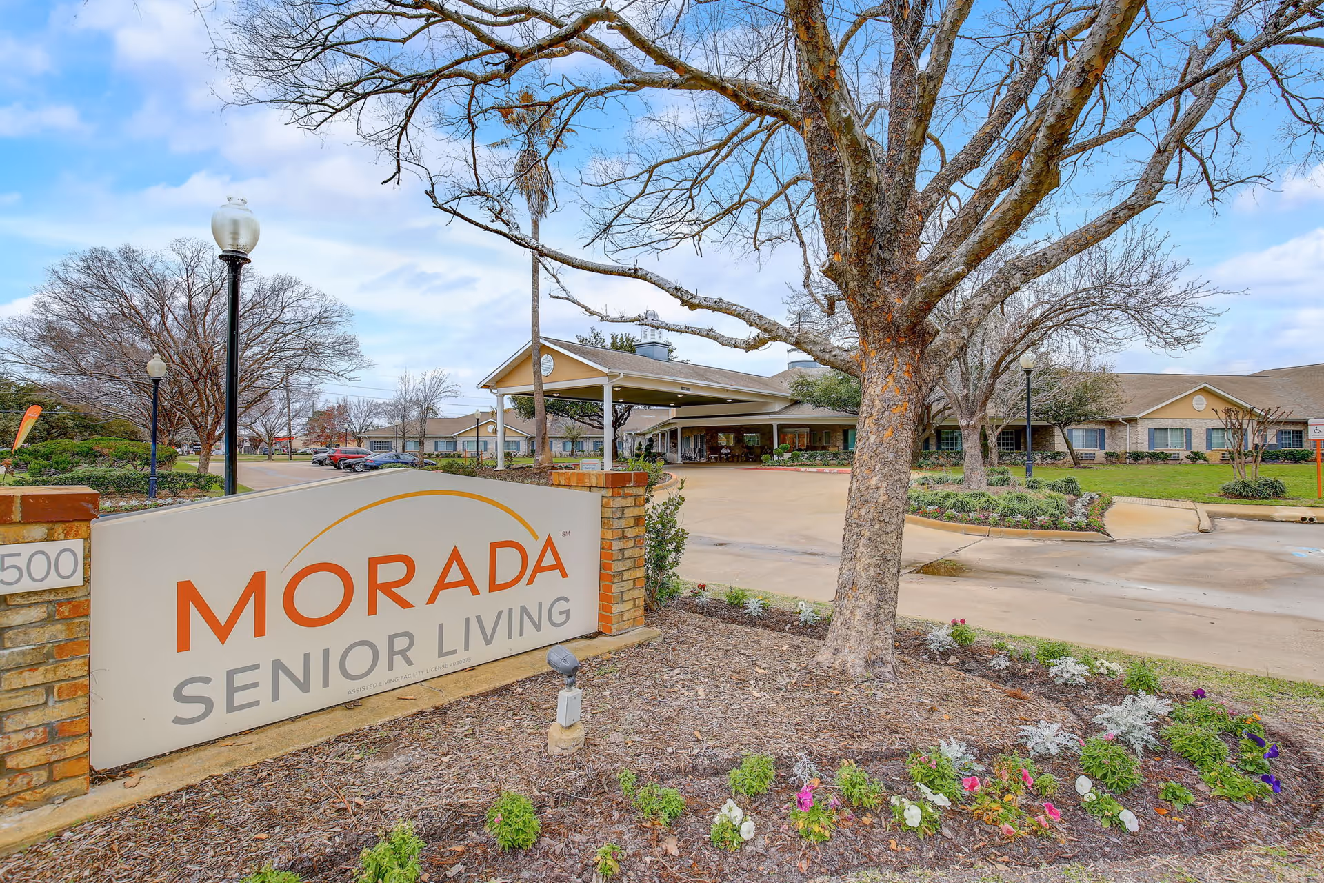 Entrance to Morada Senior Living facility with a large sign displaying the name. The building is visible in the background with a covered driveway, surrounded by landscaped grounds with trees and flower beds under a partly cloudy sky.