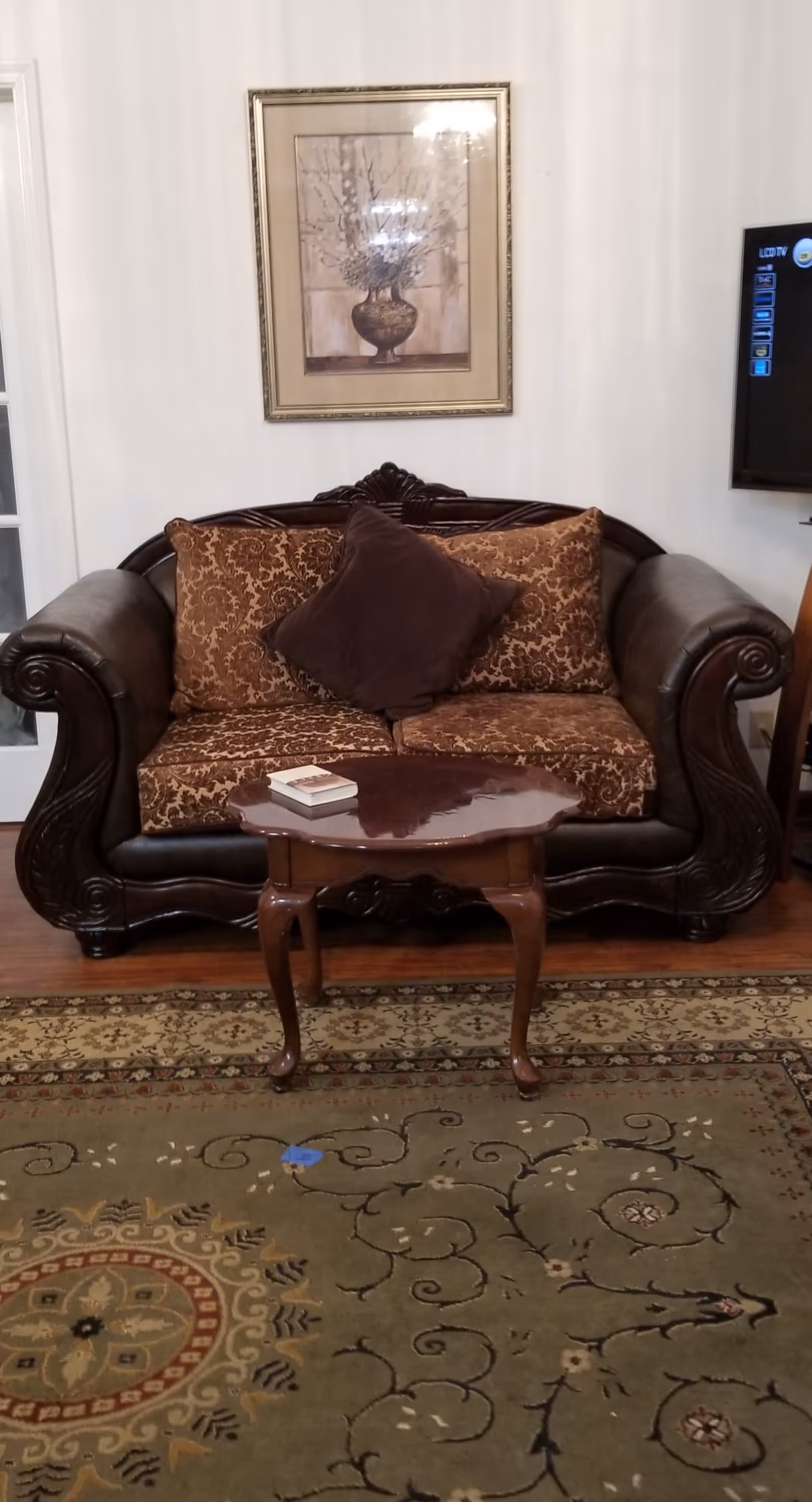A living room area featuring a dark wooden sofa with ornate carvings and brown patterned cushions, a small wooden coffee table with a book on it, a patterned area rug, a framed floral painting on the wall, and a flat-screen TV mounted on the right wall.