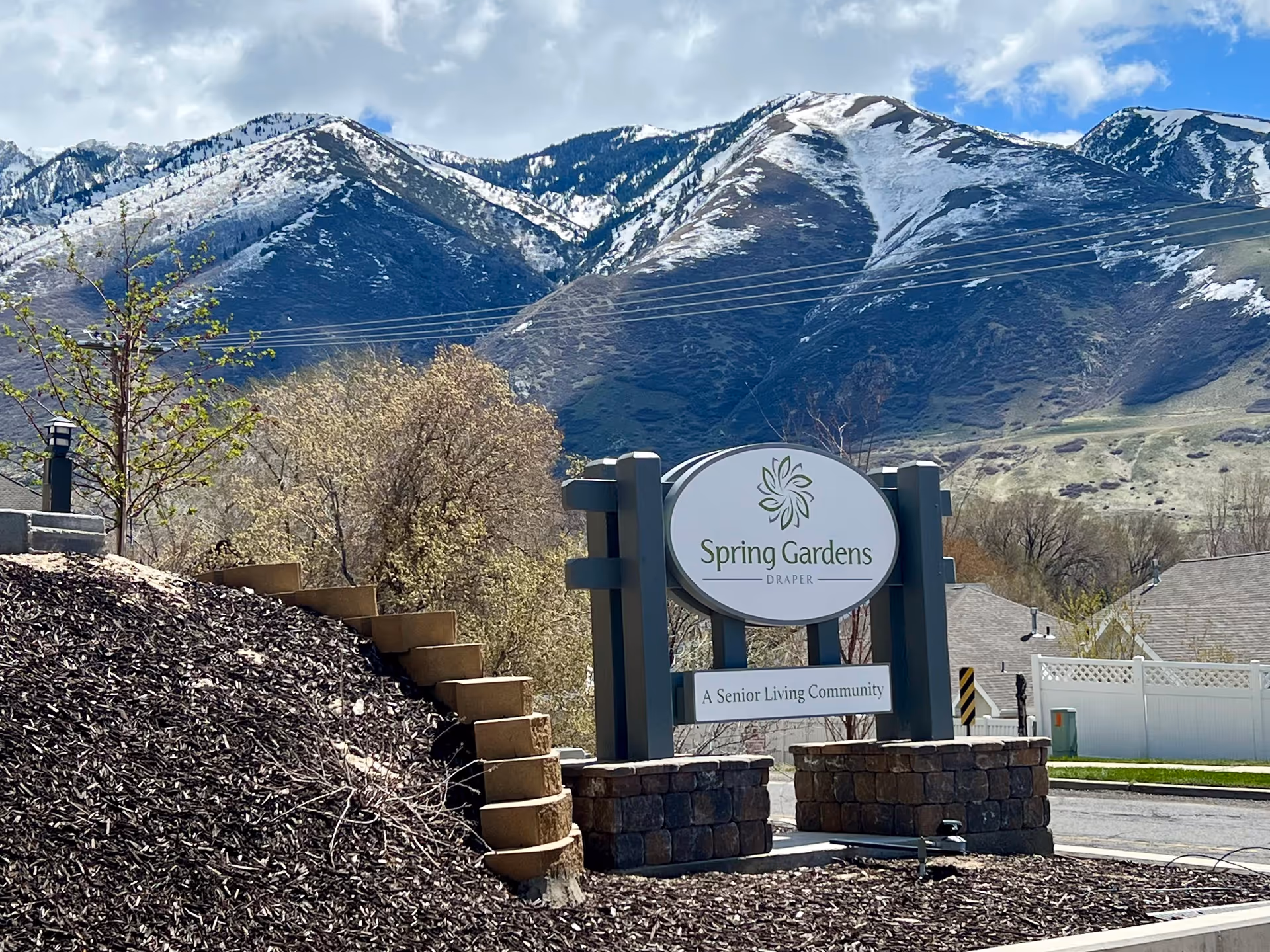 Outdoor view of the entrance sign for Spring Gardens Senior Living Draper, a senior living community, with snow-capped mountains and trees in the background under a partly cloudy sky.