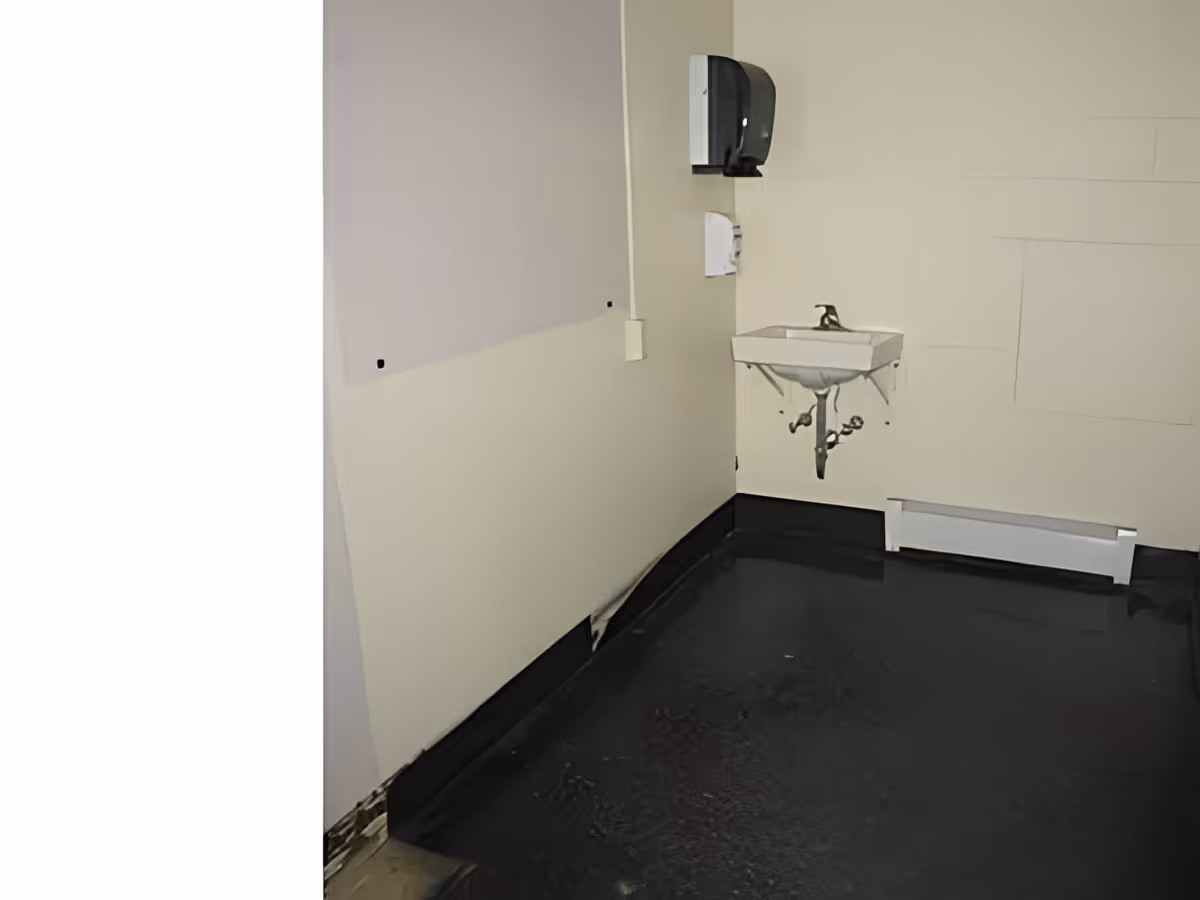 A small corner of a room with a wall-mounted white sink, a black soap dispenser above it, and a white paper towel dispenser on the wall. The floor is black and shiny, and the walls are light-colored with some visible marks and patches.