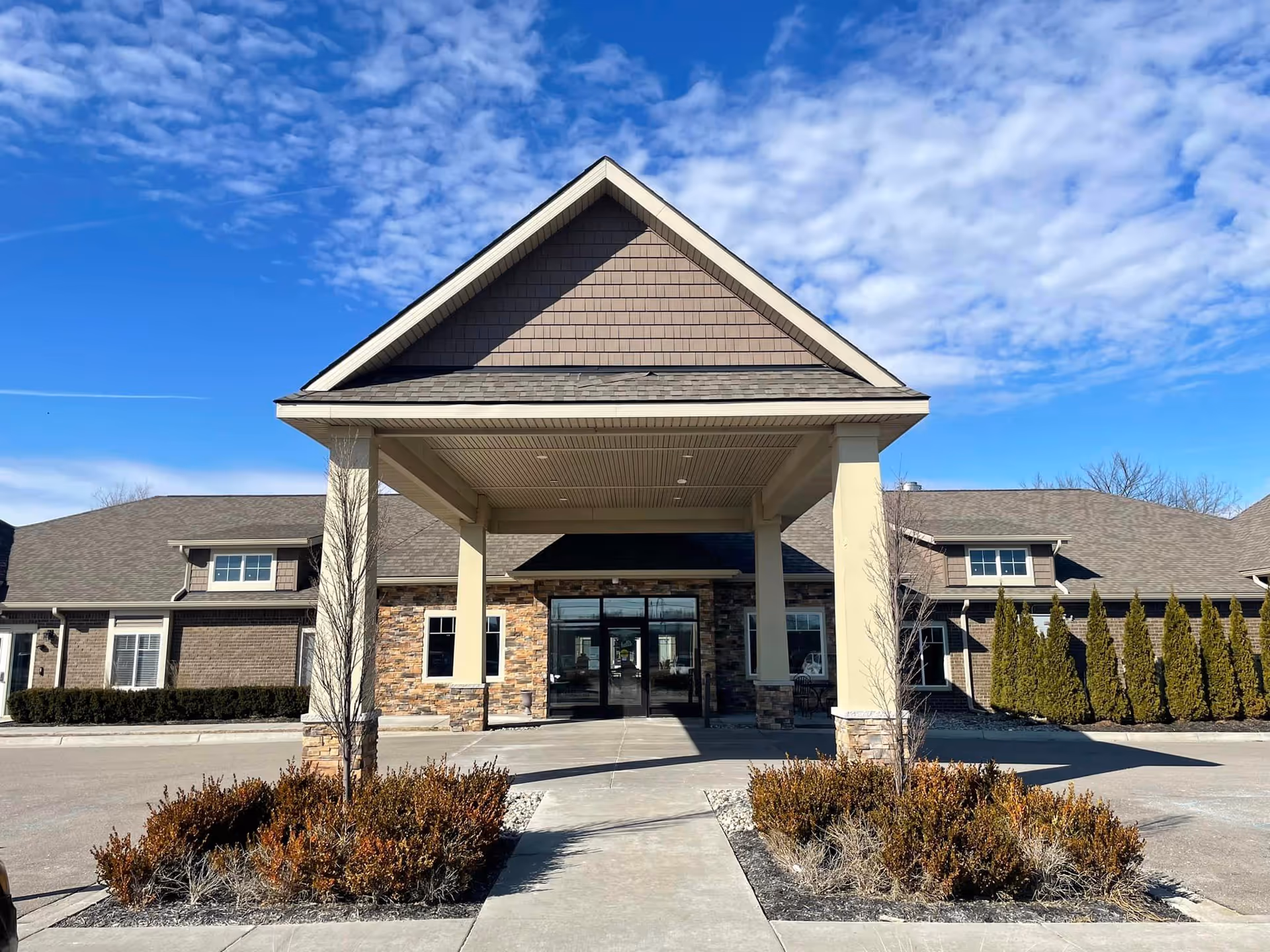 Front exterior view of Volante Senior Living of Chesterfield building with a covered entrance, stone and brick facade, and clear blue sky with scattered clouds.