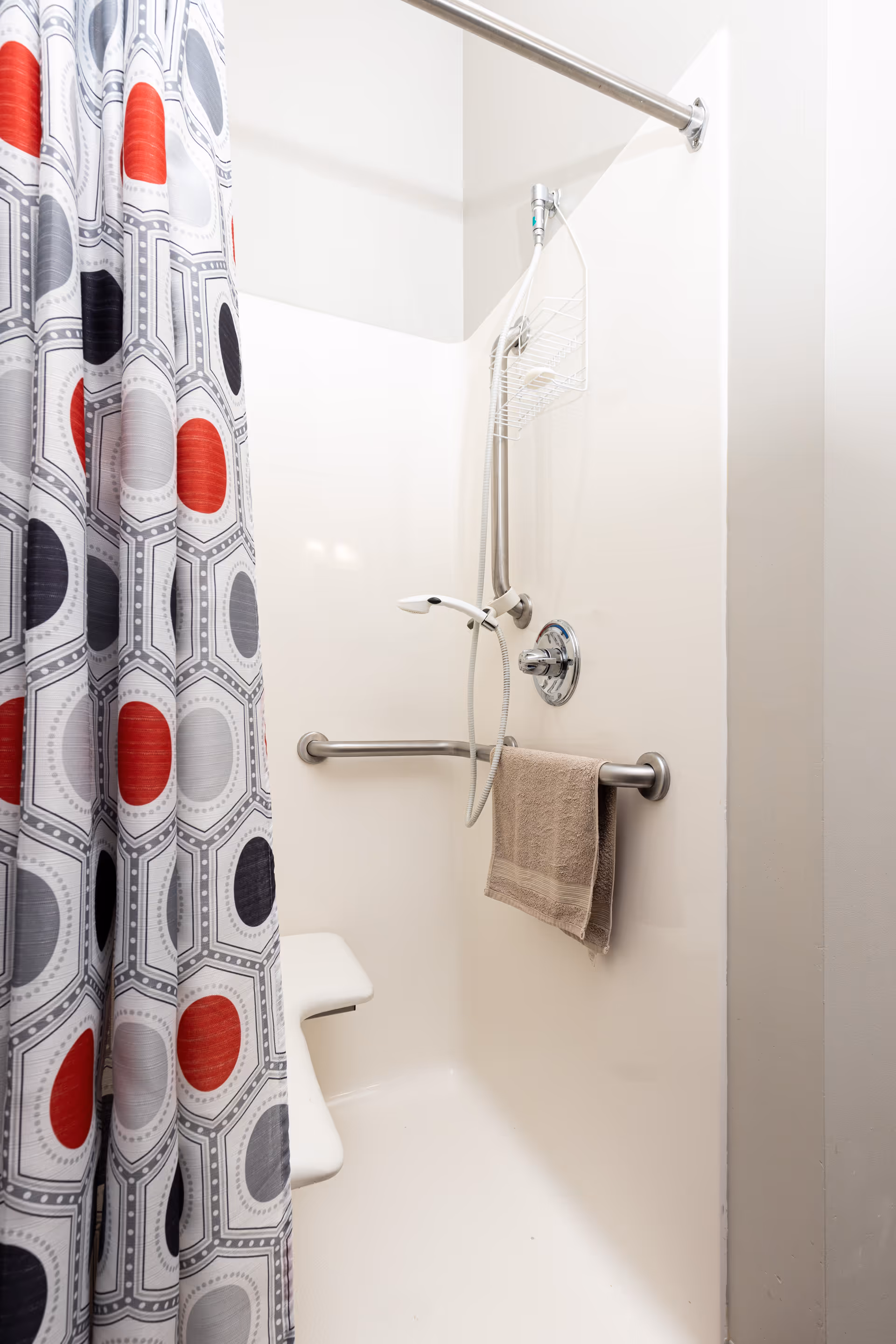 Accessible shower stall with a patterned curtain, handheld showerhead, grab bar holding a towel, and a built-in seat.