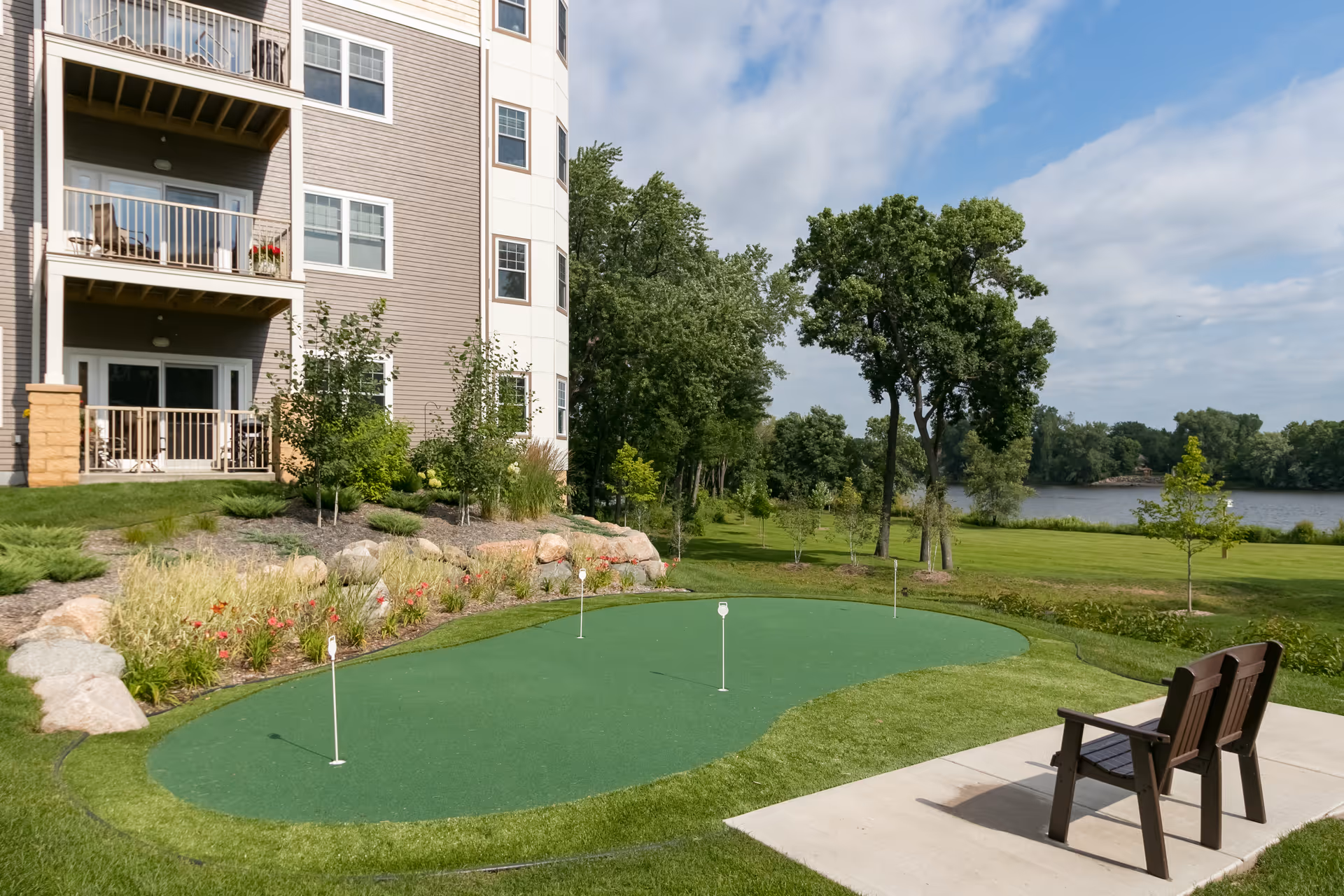 Outdoor putting green with four golf holes next to a multi-story residential building. There are two wooden chairs on a concrete patio facing the green. The area is landscaped with rocks, plants, and trees, and a river is visible in the background under a partly cloudy sky.