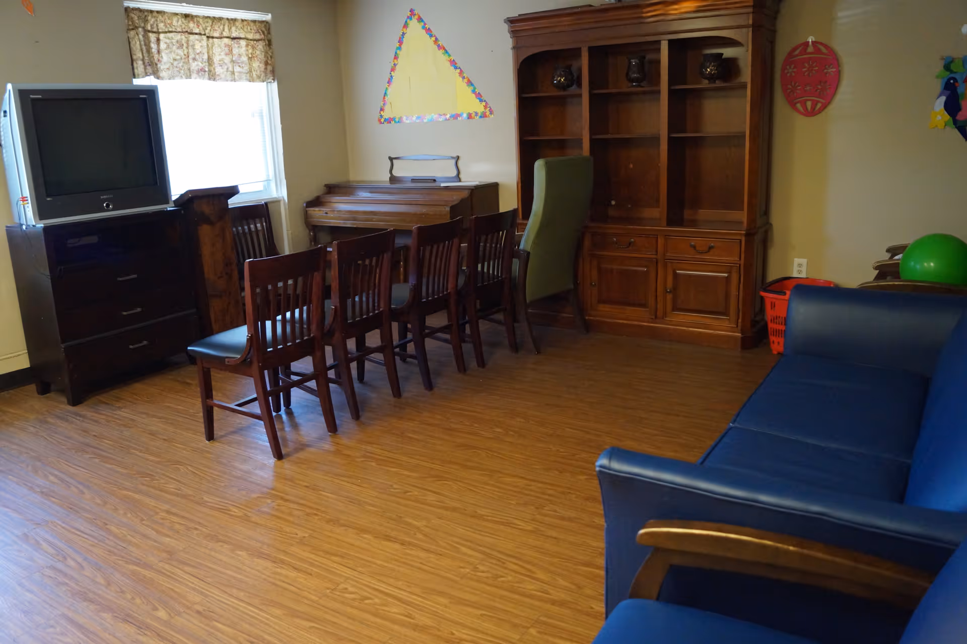 Communal sitting room with a blue couch, row of wooden chairs, TV, piano, and large wooden shelving unit.