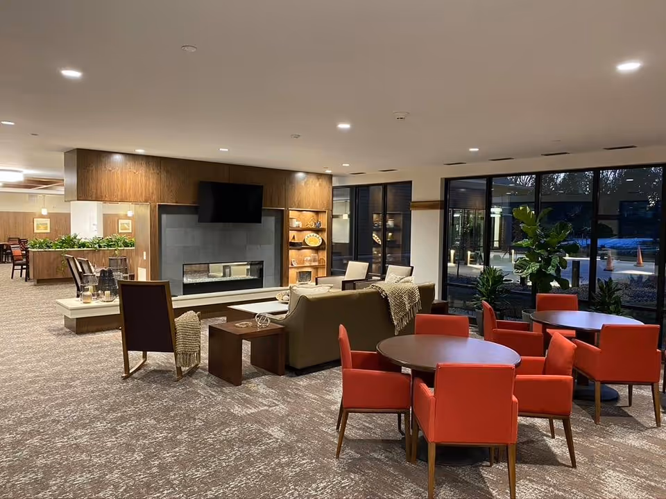 Spacious senior living lounge with sofas, a fireplace and TV, round tables with red chairs, and large windows overlooking the exterior.