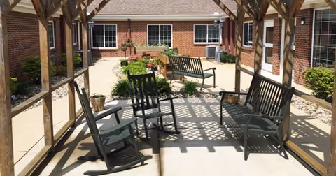 Outdoor patio area at RidgeCrest Health Campus with wooden pergola overhead, several rocking chairs and benches arranged on a concrete floor, surrounded by brick building walls and windows, with plants and landscaping along the edges.