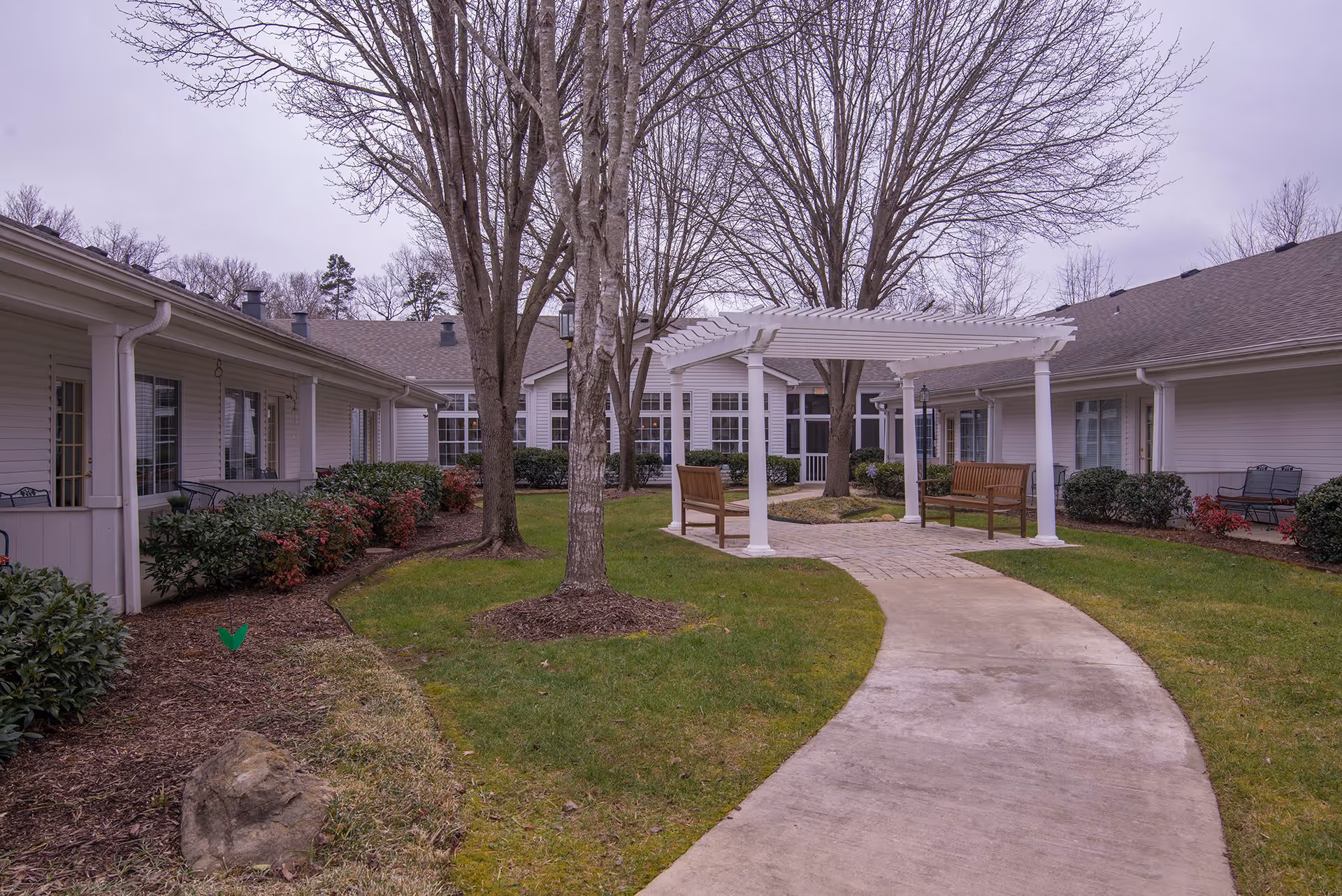 Outdoor courtyard area at Morning Pointe of Chattanooga at Shallowford featuring a curved concrete pathway leading to a white pergola with two wooden benches underneath. The courtyard is surrounded by single-story buildings with white siding and multiple windows. Leafless trees and neatly trimmed bushes and grass are visible throughout the area.