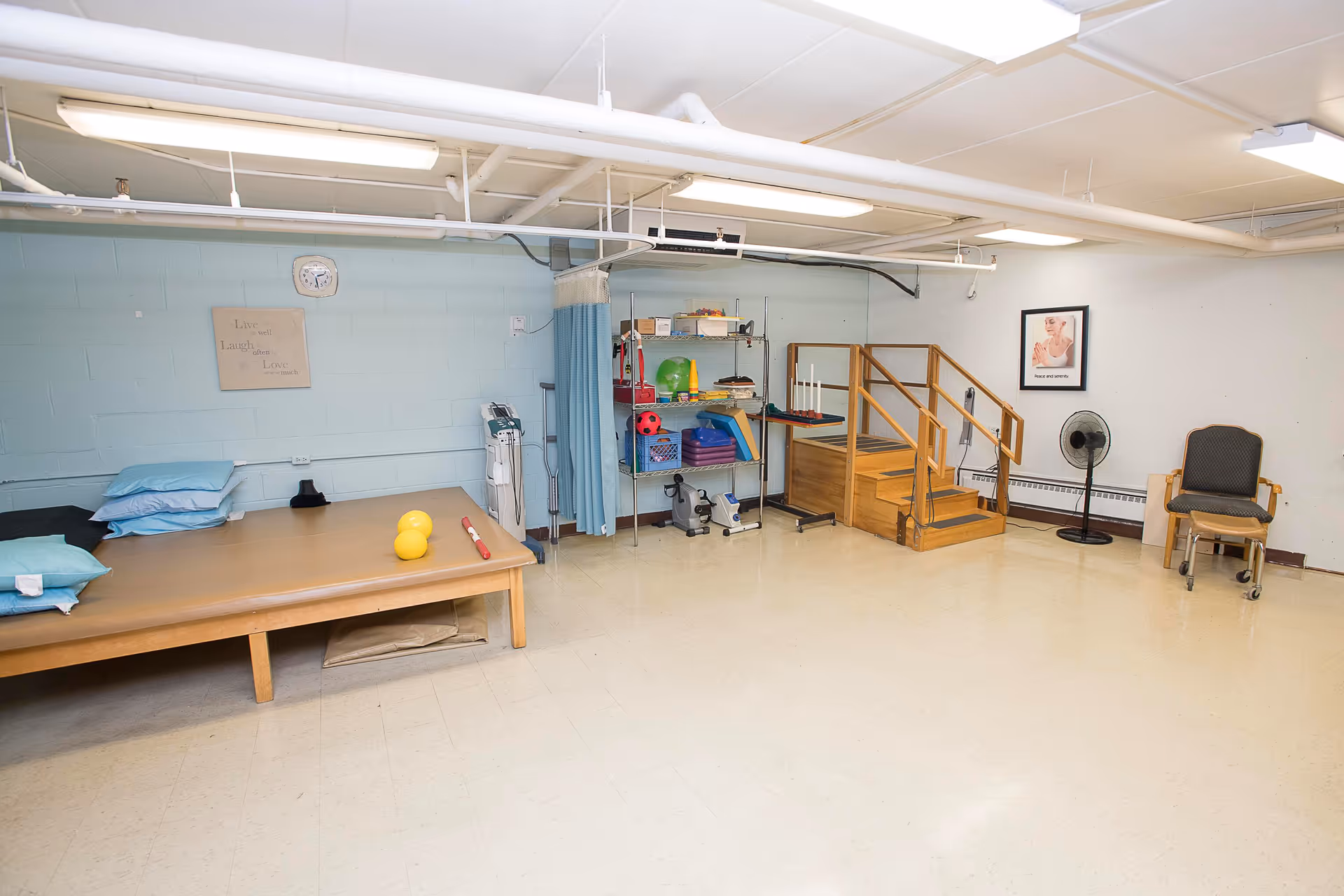 A spacious room with light-colored flooring and walls, featuring a wooden therapy table with pillows and exercise equipment including yellow weights and a red stick. There is a metal shelving unit with various therapy tools, a set of wooden stairs with handrails, a standing fan, and a chair with wheels. The room has fluorescent ceiling lights and a clock on the wall above a sign that reads 'Live Laugh Love'.