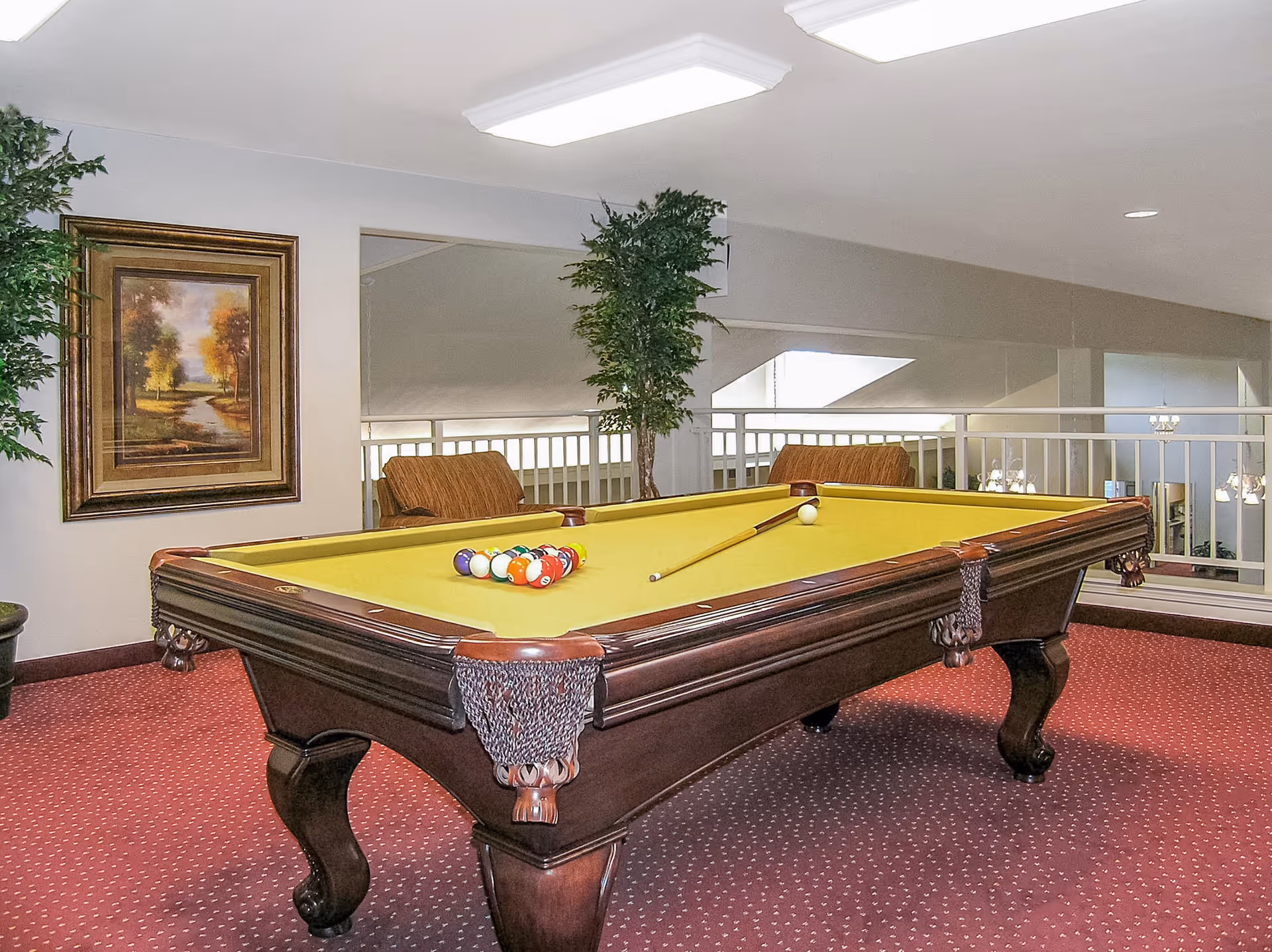A well-lit indoor recreational room featuring a yellow-felt pool table with a full set of billiard balls and a cue stick. The room has red carpet flooring, two brown cushioned chairs, a framed landscape painting on the wall, and two tall green potted plants. The background shows a white railing overlooking a lower level with chandeliers.