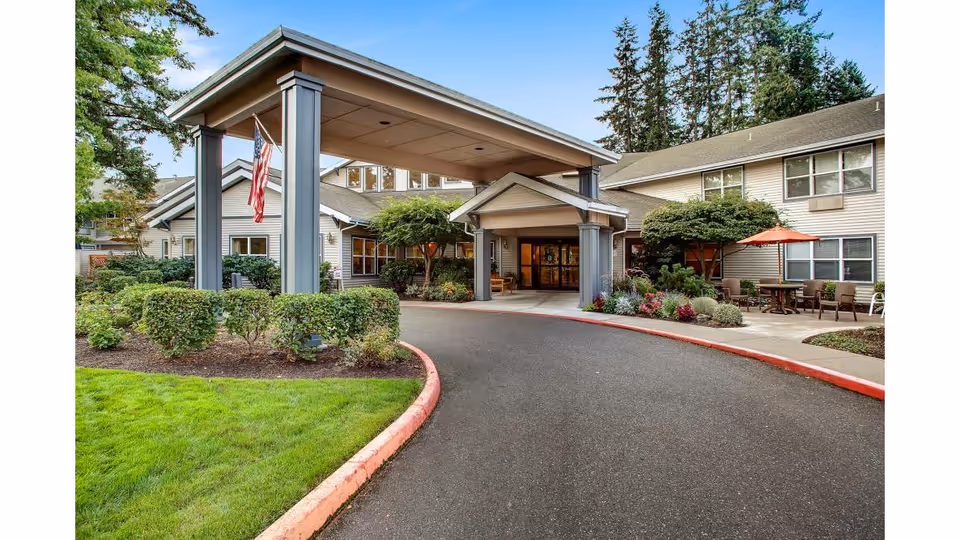 Entrance of Canfield Place senior living facility with a covered drop-off area, landscaped bushes, green lawn, and outdoor seating with an umbrella. The building is two stories with beige siding and surrounded by tall trees.