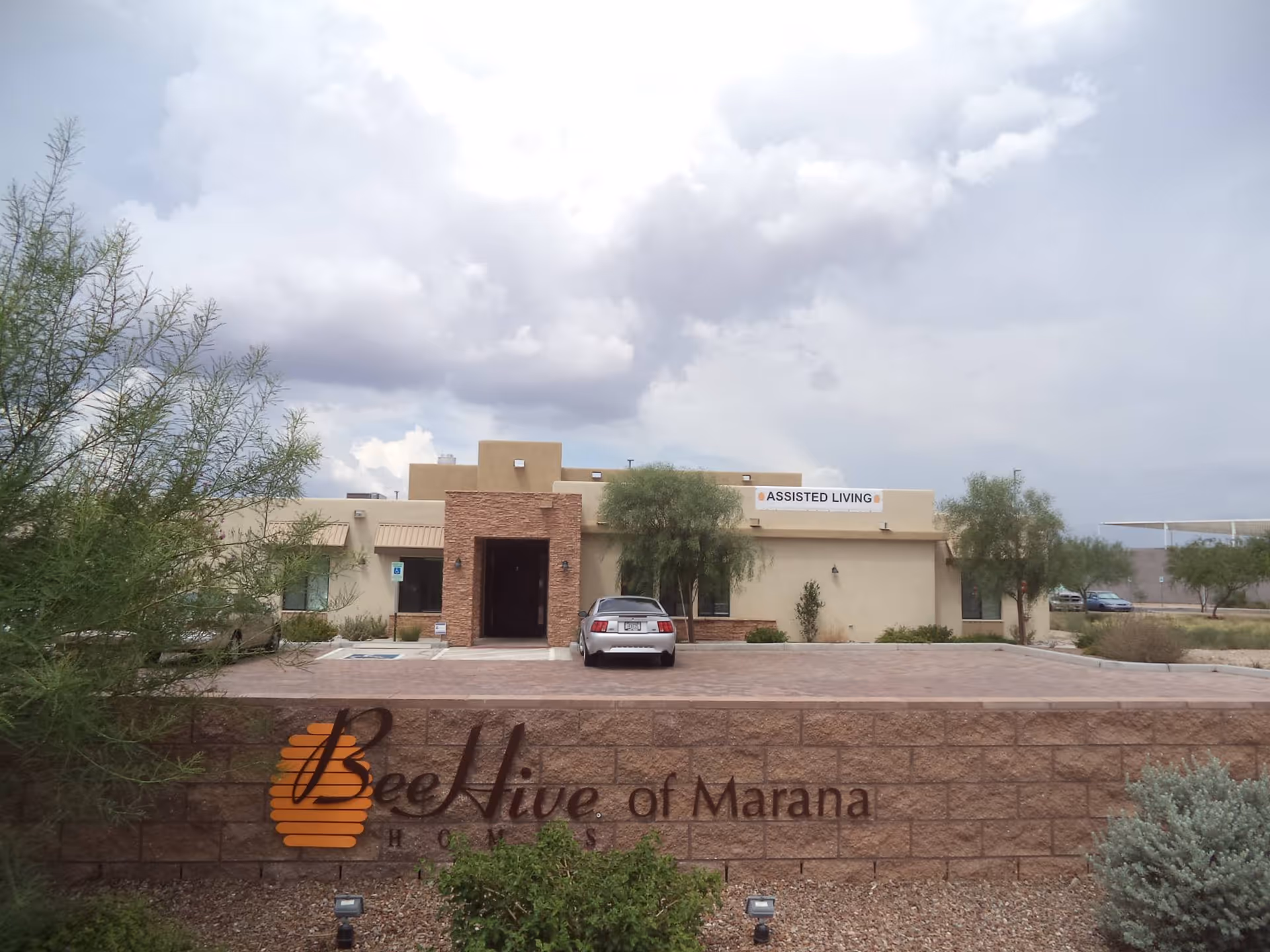 Exterior view of BeeHive Homes of Marana, a single-story assisted living facility with a beige and brown facade, a car parked in front, and a sign on the building that reads 'ASSISTED LIVING'. The foreground shows a brick wall with the facility's name and logo, surrounded by desert landscaping and trees under a cloudy sky.
