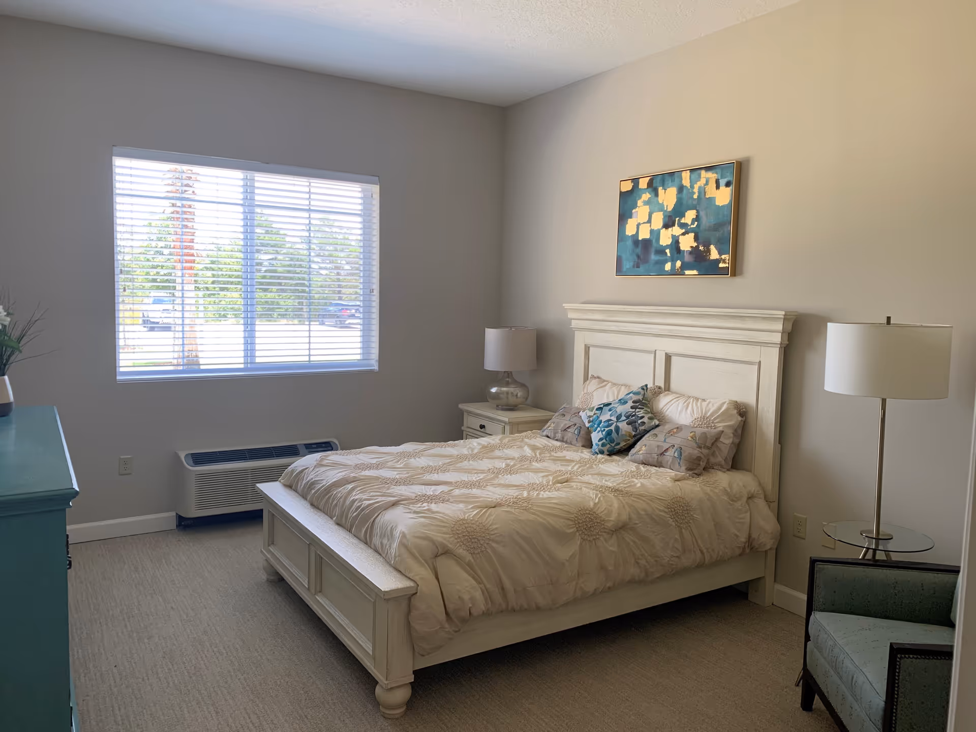 A bedroom with a large bed featuring a beige comforter and decorative pillows. There is a white wooden headboard and a matching nightstand with a lamp on the right side of the bed. A floor lamp and an upholstered chair are also visible on the right side. A window with blinds lets in natural light, and a teal dresser with a plant on top is on the left side of the room. A colorful abstract painting hangs above the bed.