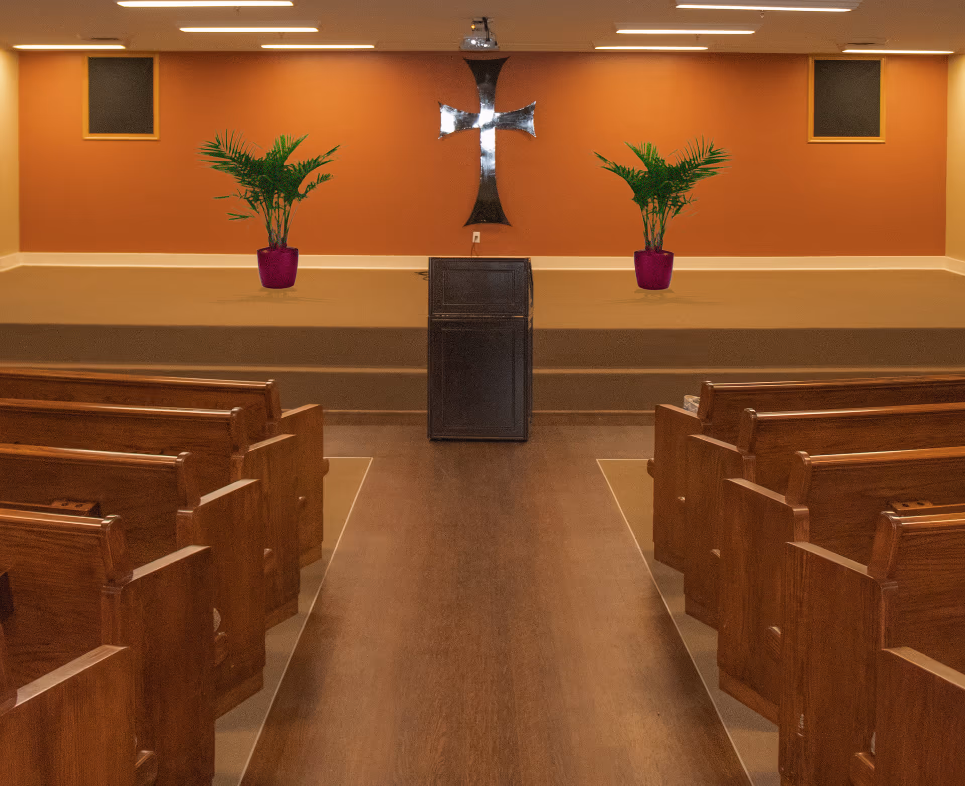 Interior chapel-style room with wooden pews, a center aisle leading to a lectern, a wall-mounted cross and two potted plants.